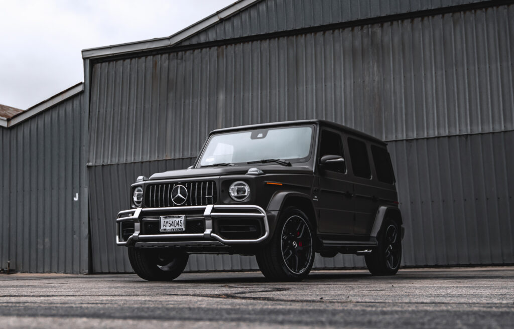 A black Mercedes Benz G 63 AMG is looking sharp as it stands ready outside a black airplane hanger on an overcast day. 