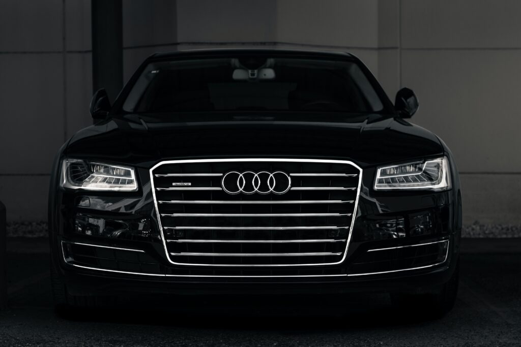 The iconic striped silver grill and interlocking circle logos of a sleek black Audi A8 Security armored car "pops" against the dark grey rectangular panels of the showroom walls.  