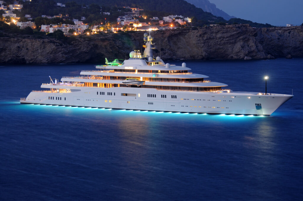 The 3-deck white-hulled superyacht, Eclipse, is anchored off a cliffy beach in the Balearic Islands of Ibiza Spain. Night is falling, and a sleepy town twinkles atop the cliffs, miles away. Eclipse herself puts on a dazzling light show with dozens of underwater circular lights, giving the stately yacht a glowing turquoise halo around it's waterline.