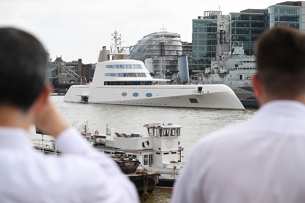 Two men in white shirts look at futuristic looking white yacht with wrap around windows above the main deck, moored on the Thames in London England. 