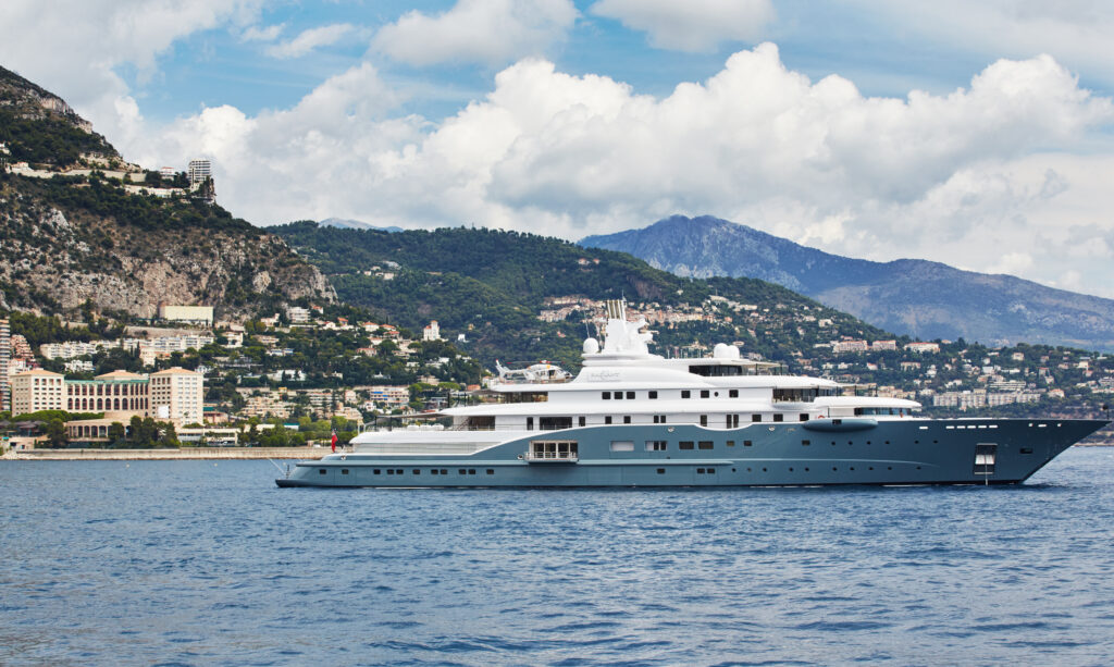 The Radiant, superyacht with a ocean-blue lower hull and white upper decks, is spotted sailing out to sea near the hilly coast of Monte Carlo.