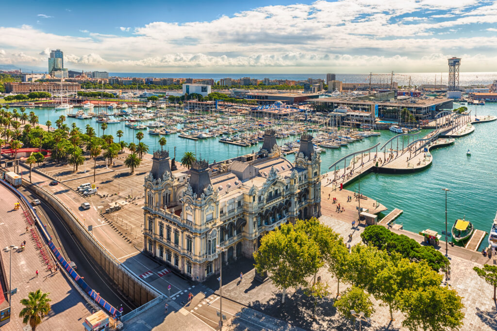 Scenic aerial view of Port Vell from the top of Columbus Monument, Barcelona, Catalonia, Spain. A modern curving pedestrian bridge extends out from historic building in Barcelona and across the aquamarine waters of Port Vell, hemming in numerousdocked ships and yachts to the left. 