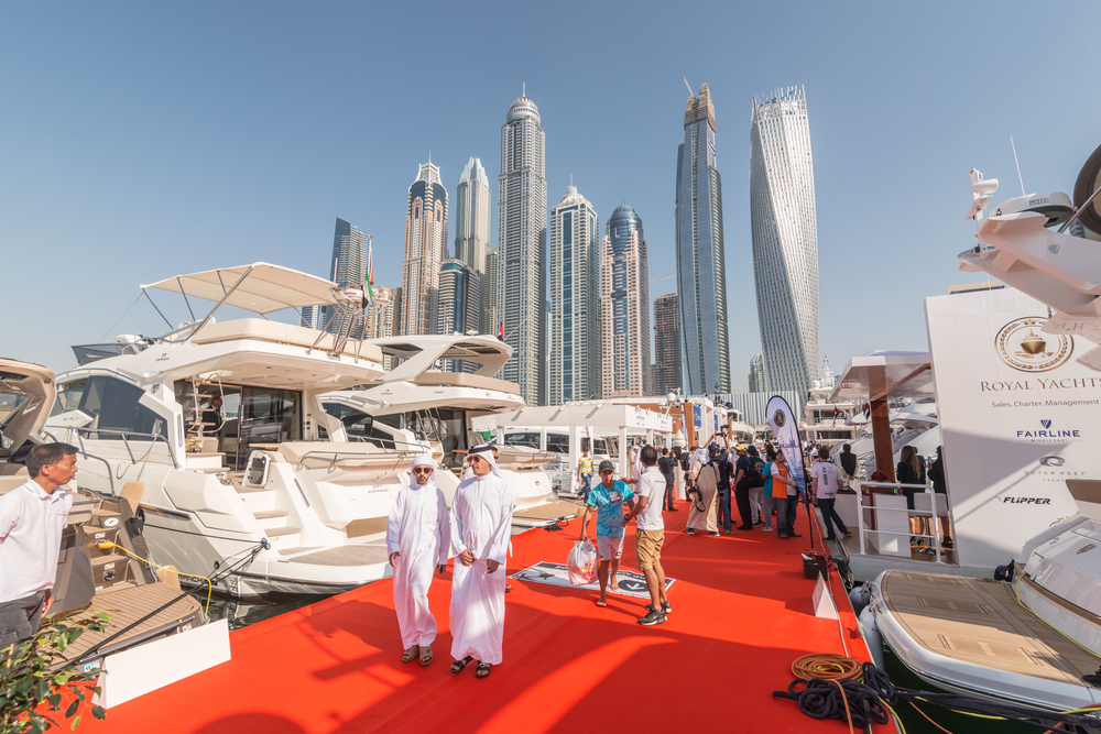 Men wearing white traditional kufiyah head coverings walk along a dock that has been converted to a red carpet showcasing yachts on either side as part of the Dubai Boat Show. In the background the Dubai Skyline dwarfs a crowd of tourists at the docks below. 