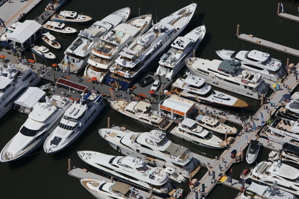 An aerial shot of large white yachts gleaming in the midday sun against a dark blue sea at the port of Palm Beach. Many passers-by hustle between the sleek ships along the grey L shaped pier. As part of the Palm Beach International Boat Festival festivities, solid red beach umbrellas and matching floor mats seem to mark entrances to participating vessels. Two luxury sedans— one black, the other white— have been driven on to a floating platform and docked among the yachts.