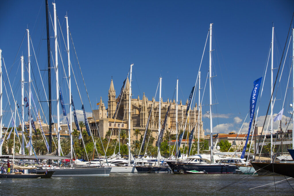 At a calm marina, classic sailing yachts in blue and white rest on their moorings, their masts gracing the sky. Through this veritable forest of ships masts, an old Spanish style gothic cathedral can be seen on a nearby hill.