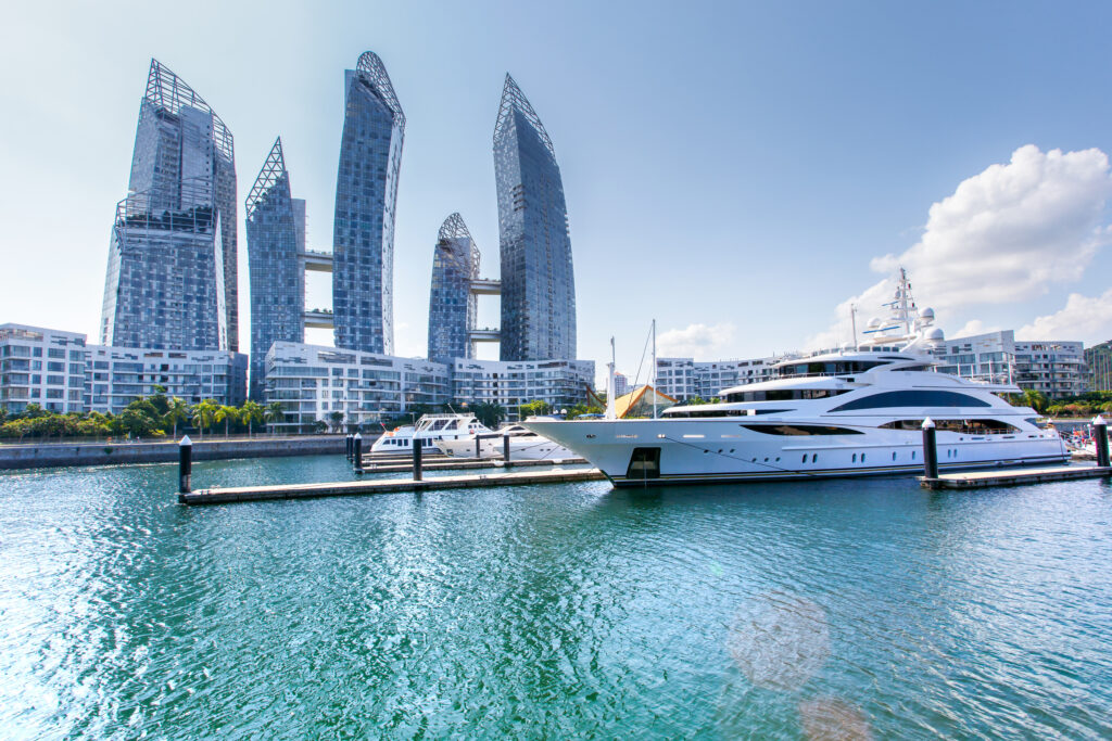 A fancy white yacht is docked in the turquoise waters of Singapore near some futuristic angled buildings with curving glass facades. 