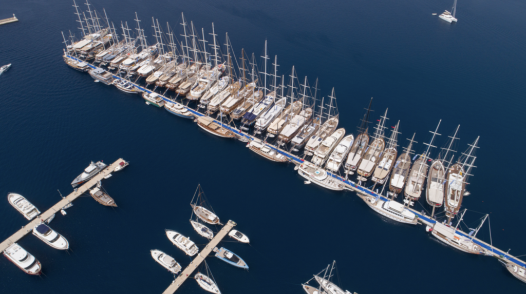 An aerial view of some ships docked at TYBA on sapphire hued seas that are glass-like with calm. Blue carpeting has been rolled out over the docks for this special event. On one side of these docks, traditional wooden sailing ships rest side by side. On the other side, a mix of modern and traditional ships are docked stem to stern. 
